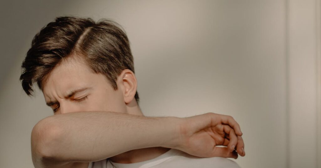 A young man sneezes into his elbow, showcasing a hygiene practice, in an indoor setting.