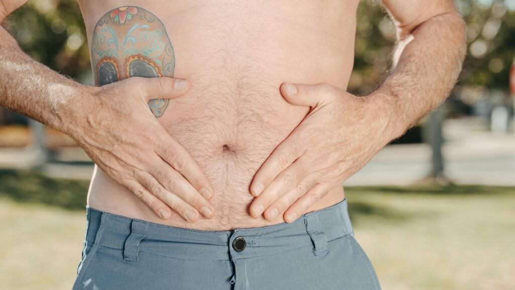 A man with a tattoo on his torso holds his stomach outdoors, suggesting discomfort.